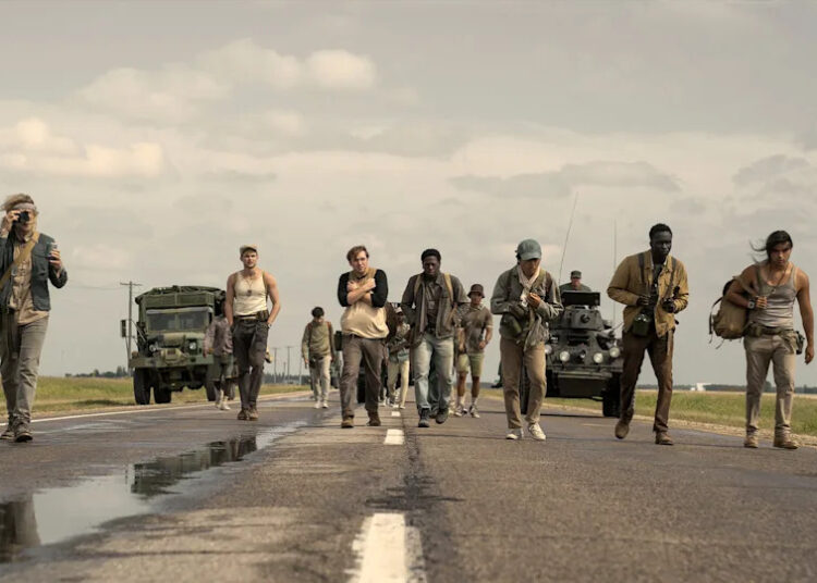 A group of people walking down a rural road, with military trucks and tanks in the background under a cloudy sky.