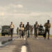 A group of people walking down a rural road, with military trucks and tanks in the background under a cloudy sky.