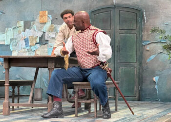 Two men performing on stage during a play. One has an enhanced and disfigured nose and is sitting at a bench, next to a table, with his arms outstretched as he speaks to another man over his shoulder.