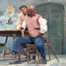 Two men performing on stage during a play. One has an enhanced and disfigured nose and is sitting at a bench, next to a table, with his arms outstretched as he speaks to another man over his shoulder.