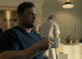 Alan Ritchson's Jack Reacher holds a coffee mug as he sits at a kitchen table in Reacher