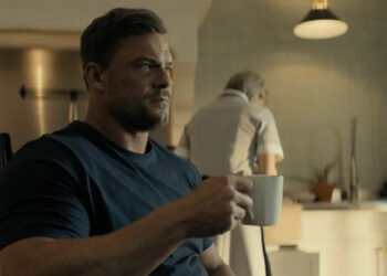 Alan Ritchson's Jack Reacher holds a coffee mug as he sits at a kitchen table in Reacher