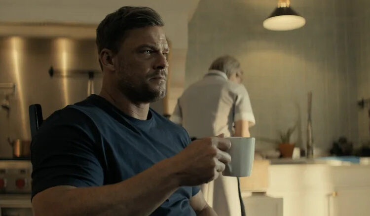 Alan Ritchson's Jack Reacher holds a coffee mug as he sits at a kitchen table in Reacher