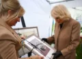 PA Media Camilla wears a brown jacket with a glittery broach and holds a portrait of a small dog. Another woman in a brown jacket also holds the portrait.