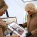 PA Media Camilla wears a brown jacket with a glittery broach and holds a portrait of a small dog. Another woman in a brown jacket also holds the portrait.