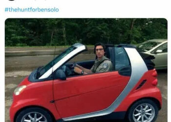 Adam Driver in a red Smart Car staring at the camera seriously.