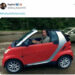 Adam Driver in a red Smart Car staring at the camera seriously.