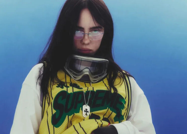 Billie Eilish wearing a Supreme jersey and goggles, with long dark hair and a serious expression against a blue background.