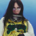Billie Eilish wearing a Supreme jersey and goggles, with long dark hair and a serious expression against a blue background.