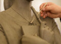 A close-up of a sculpture of a woman being crafted, focusing on the neck, shoulders and chest. The sculpture is clutching a book in their hands across the chest. The sculptor is using a fine stick to craft the sculpture.