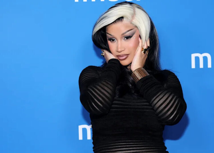 Cardi B with black and white hair poses against a blue background wearing a black outfit.