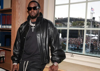 A man in a black leather jacket and sunglasses stands by a window overlooking a large outdoor event with a stage and crowd.