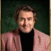 Jonathan Ross in the show's official portrait, smiling and wearing a black top and salmon coloured blazer
