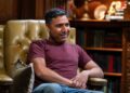 Nick Mohammed wearing a red top and blue jeans, smiling ahead.