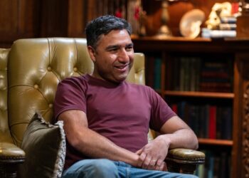 Nick Mohammed wearing a red top and blue jeans, smiling ahead.