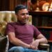 Nick Mohammed wearing a red top and blue jeans, smiling ahead.