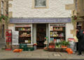 A painted white cotswold stone building with hanging baskets on either side. the bottom floor is a shop front, with Cotchester Convenience Store on it. It has had 'Proud to stock the Rutshire Herald' on the window and outside are baskets of fruit and veg, 1980s sweet machines and a rack with newspapers.