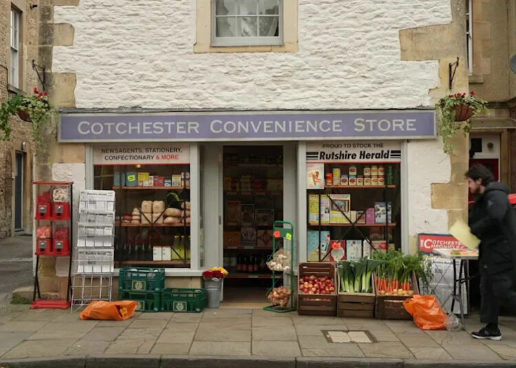 A painted white cotswold stone building with hanging baskets on either side. the bottom floor is a shop front, with Cotchester Convenience Store on it. It has had 'Proud to stock the Rutshire Herald' on the window and outside are baskets of fruit and veg, 1980s sweet machines and a rack with newspapers.