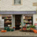 A painted white cotswold stone building with hanging baskets on either side. the bottom floor is a shop front, with Cotchester Convenience Store on it. It has had 'Proud to stock the Rutshire Herald' on the window and outside are baskets of fruit and veg, 1980s sweet machines and a rack with newspapers.