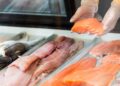 A grocery store employee's hands hold a piece of salmon inside a display case for seafood.