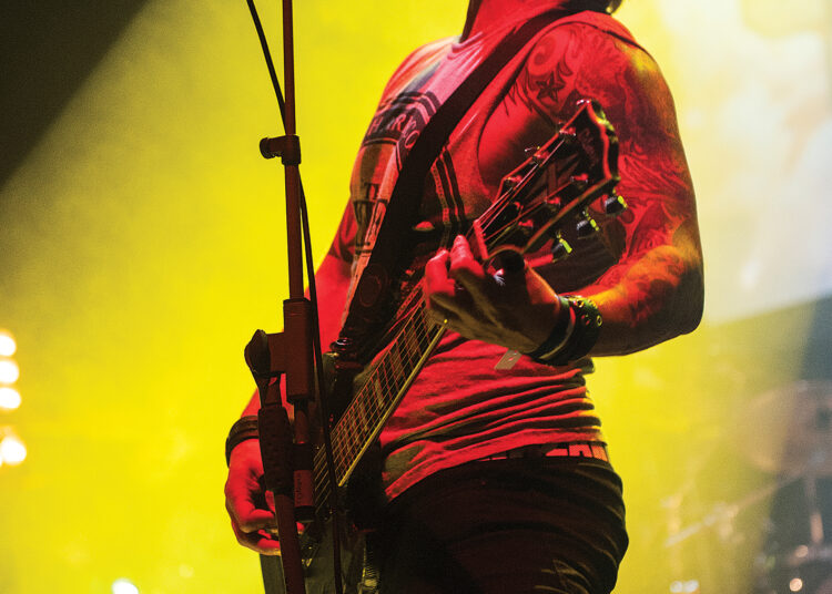 A guitarist passionately performs on stage, illuminated by vibrant yellow lighting, showcasing intricate tattoos and an electric guitar