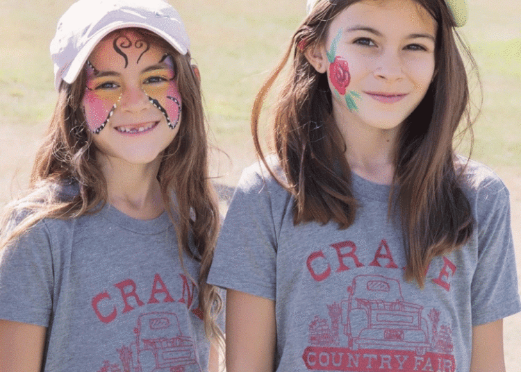 Two girls wearing white baseball caps and gray Crane t-shirts, show off their face paintings that are always a hit at the Crane Country Fair. (Courtesy photo)