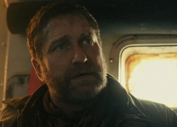 A bearded man (Gerard Butler) looking worried, stood in front a small window.