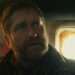 A bearded man (Gerard Butler) looking worried, stood in front a small window.