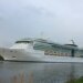 A large white cruise ship, named "Serenade of the Seas," is sailing on a calm body of water with a cloudy sky in the background as it makes its way toward Amsterdam.