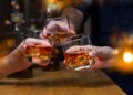 Three hands holding glasses of whiskey with ice, clinking them together.
