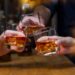 Three hands holding glasses of whiskey with ice, clinking them together.
