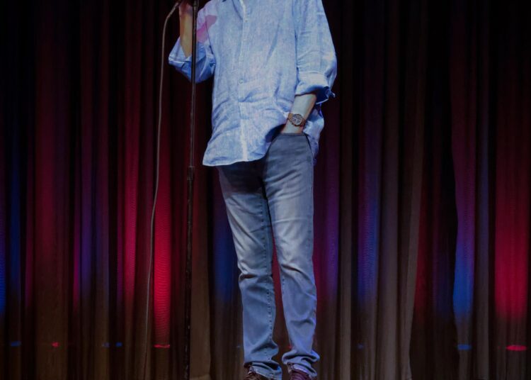 A man in blue shirt and jeans stands at a mic stand on stage.