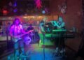 Members of a band on a colorfully lit stage playing flute, guitars, drums, and bass guitar.