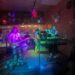 Members of a band on a colorfully lit stage playing flute, guitars, drums, and bass guitar.