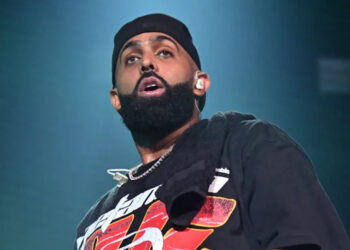 Eladio Carríon with a beard and black cap performing on stage, wearing a graphic t-shirt and a towel around his neck.