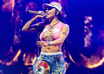 GloRilla performing on stage, wearing a pink top and decorated jeans, holding a microphone. The background is lit with colorful lights.