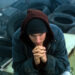Eminem in a hoodie and beanie sits pensively among piles of tires in an industrial setting.