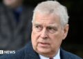 Prince Andrew wearing a dark textured overcoat over a white shirt and red tie, standing outdoors with a blurred background