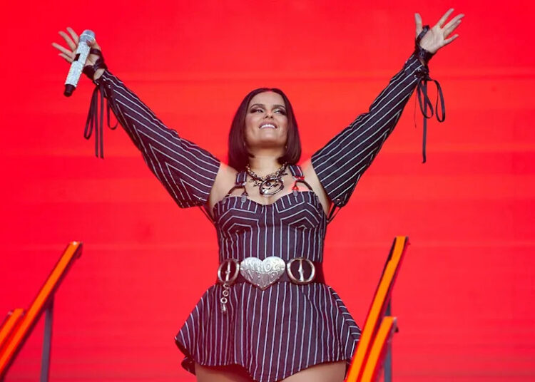 Nelly Furtado performing live on stage with her hands up to the crowd in front of a red background.
