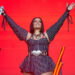 Nelly Furtado performing live on stage with her hands up to the crowd in front of a red background.