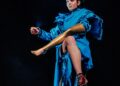 An actor in a blue gown with a flashlight mounted on her head sits on a stool while holding a gold leg.