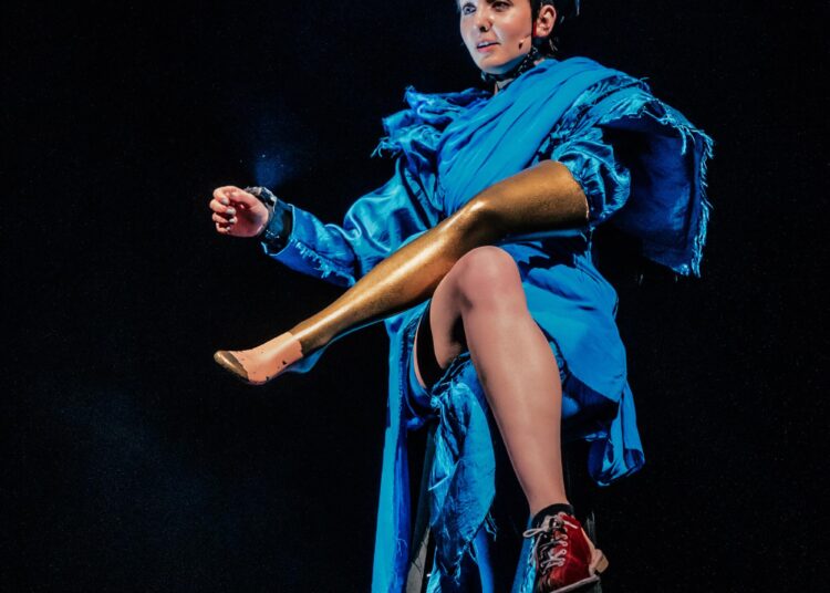 An actor in a blue gown with a flashlight mounted on her head sits on a stool while holding a gold leg.