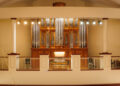A large pipe organ with ornate woodwork is displayed behind a railing in a well-lit concert hall.