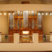 A large pipe organ with ornate woodwork is displayed behind a railing in a well-lit concert hall.
