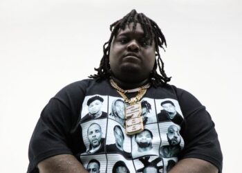 Dallas rapper HeadHuncho Amir wears a black shirt and gold chains standing in front of a white background looking straight to the camera.
