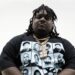 Dallas rapper HeadHuncho Amir wears a black shirt and gold chains standing in front of a white background looking straight to the camera.