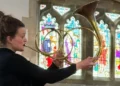 Ursula Paludan Monberg A woman plays an unusual brass horn in front of a stained glass window in a church. The horn is a golden, brassy colour and does three different loops in the metal before eventually widening to allow the sound to come out