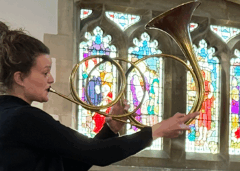 A woman plays an unusual brass horn in front of a stained glass window in a church. The horn is a golden, brassy colour and does three different loops in the metal before eventually widening to allow the sound to come out