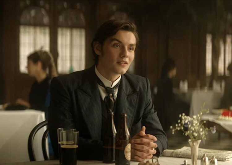 Louis Partridge in a suit, sitting at a table with two bottles of Guinness in front of him in House of Guinness.