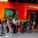 A group of people lined up on a sidewalk, behind a velvet rope, in front of a building with a bright orange facade, and the words "Taylor Swift" over the entrance. On the wall above the lined up people is an image of an album cover with a woman, Taylor Swift, laying in water with her hand behind her head.